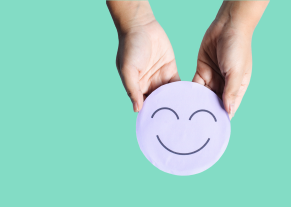 Zwei H&auml;nde halten eine lilafarbene, lachende Emoji-Gesichtscheibe vor gr&uuml;nem Hintergrund.