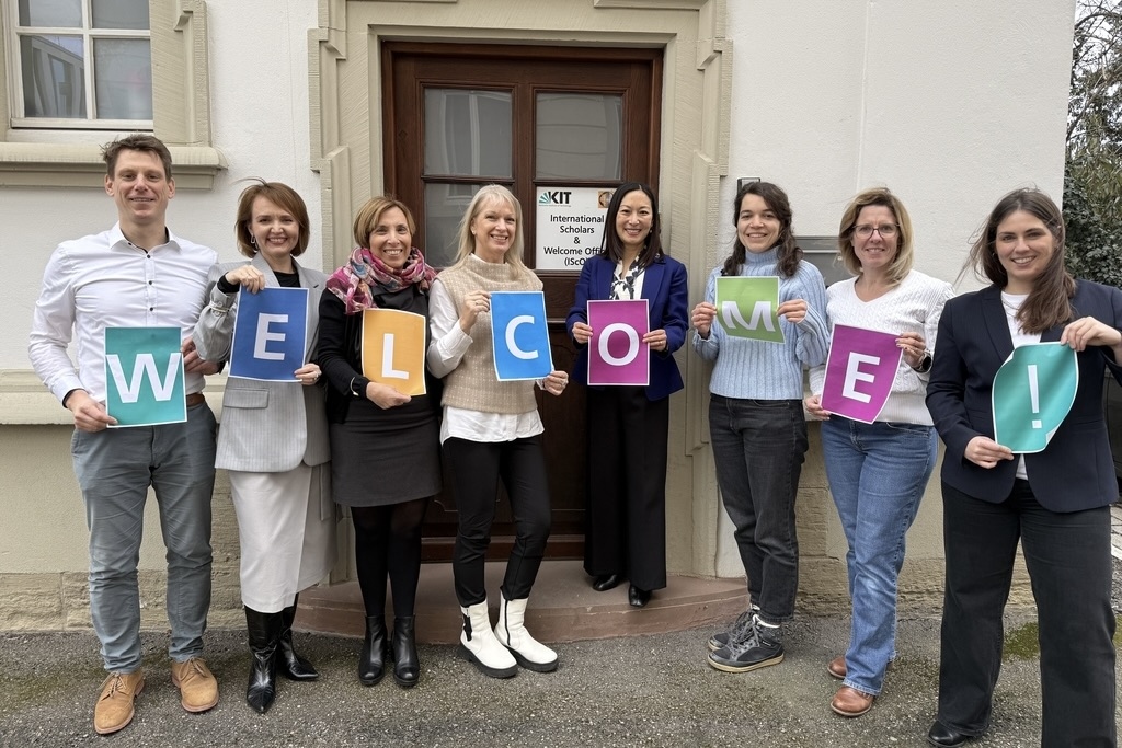 Gruppe von acht Personen vor Tür, halten farbige Buchstaben, die „WELCOME!“ ergeben.