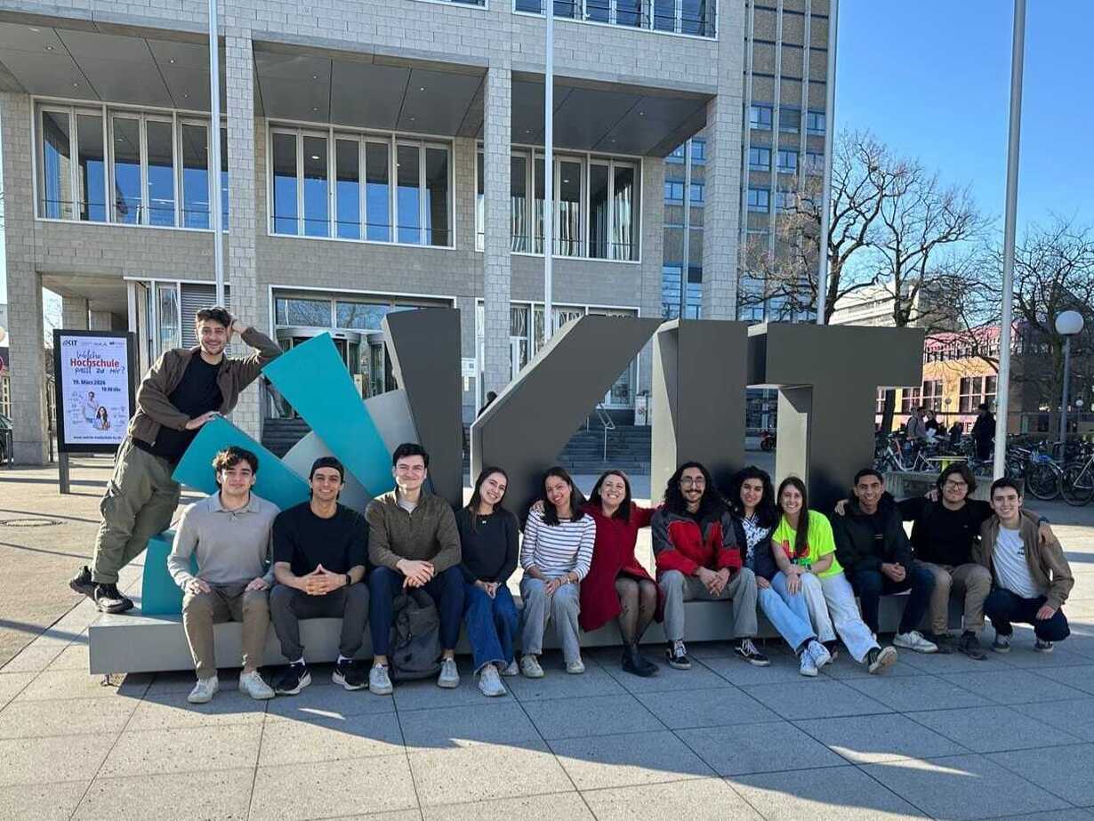 Gruppe junger Menschen posiert vor einem großen Schriftzug vor einem Universitätsgebäude.