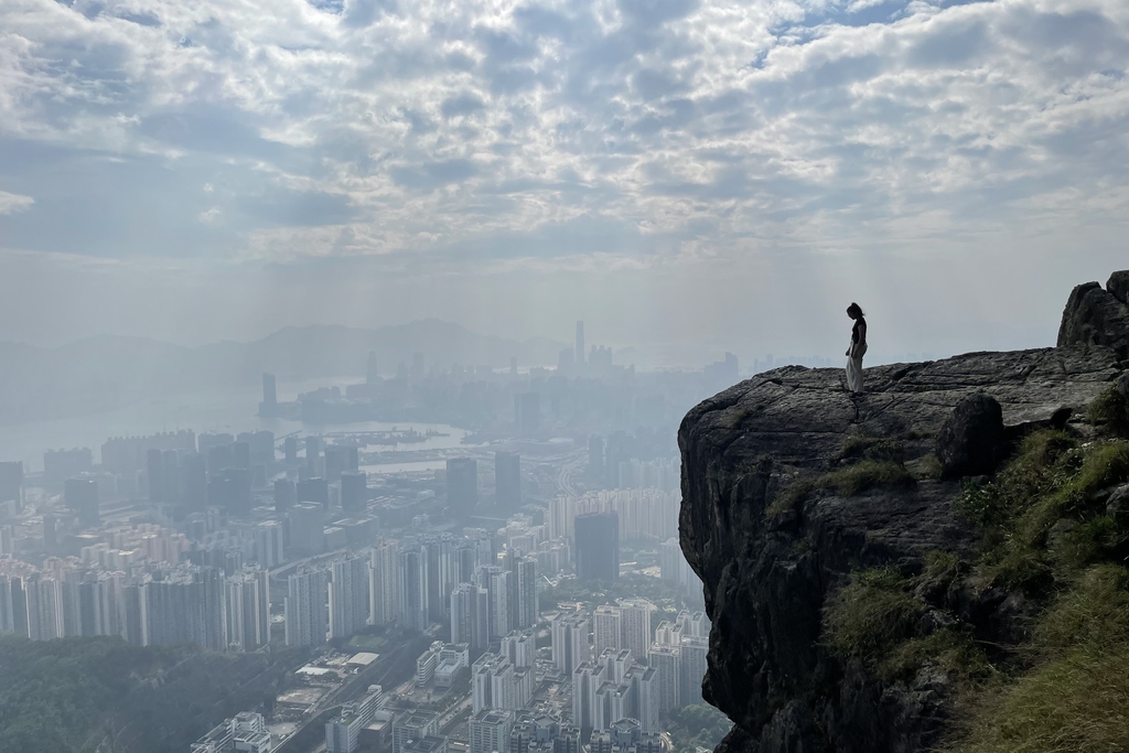 Person steht auf Felsvorsprung mit Blick auf Stadtpanorama unter bewölktem Himmel.