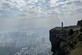 Person steht auf Felsvorsprung mit Blick auf Stadtpanorama unter bewölktem Himmel.