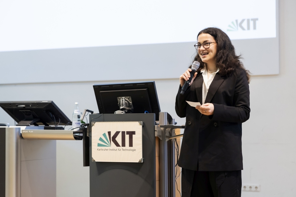 Woman speaking into a microphone at a KIT podium during a presentation.