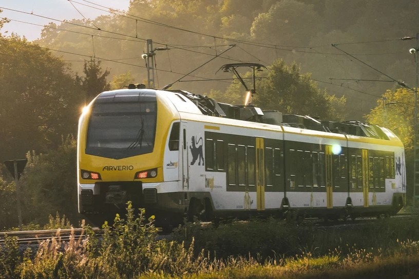 Nahverkehrszug fährt durch grüne Landschaft, umgeben von Bäumen und Oberleitungen.