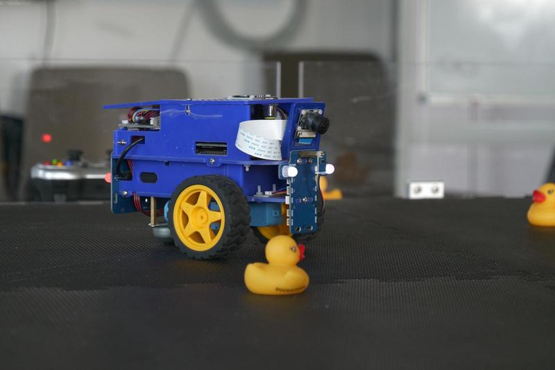 Blauer Forschungsroboter mit gelben Rädern und Gummienten auf einer schwarzen Testfläche.