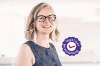 Frau mit kurzem, blondem Haar trägt blaues, ärmelloses Kleid vor hellem Hintergrund.