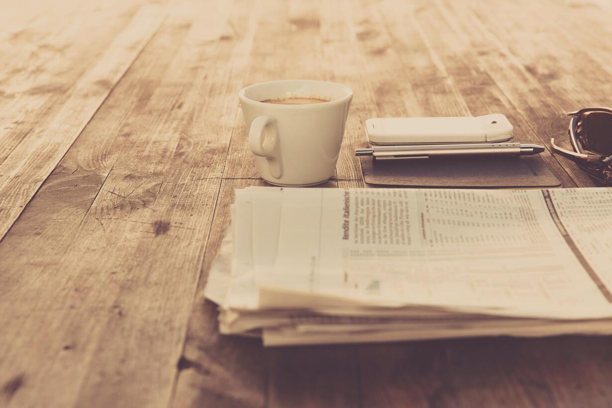 Tasse Kaffee, Zeitung, Handy und Stift auf einem Holztisch.