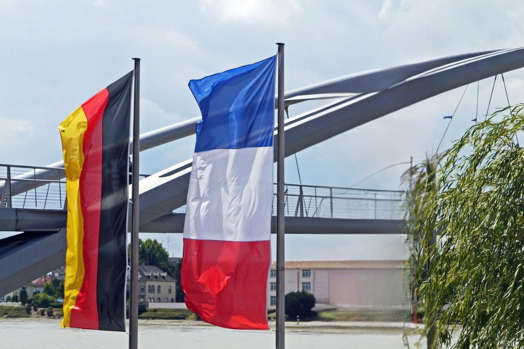 Drei Fahnen: Deutschland, Frankreich und daneben eine Brücke über einem Wasserarm.